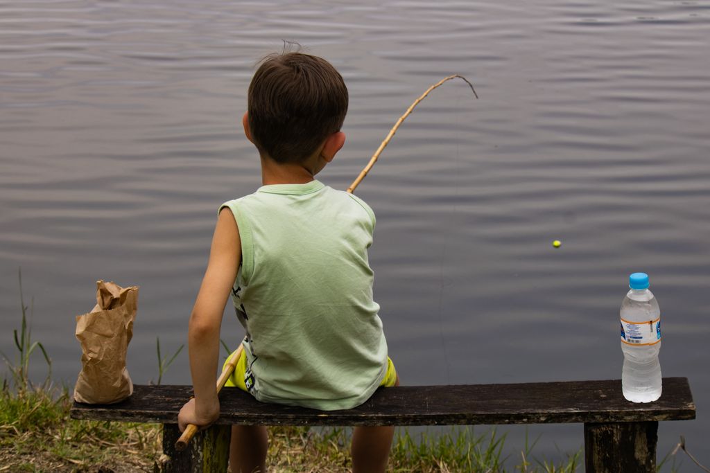 A young boy sits on a bench fishing by a serene lake under the daylight sun.